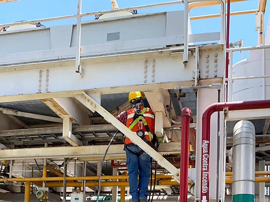 Trabajador con casco y arnés sobre la estructura de un patín de proceso, junto a la línea roja de agua contra incendio