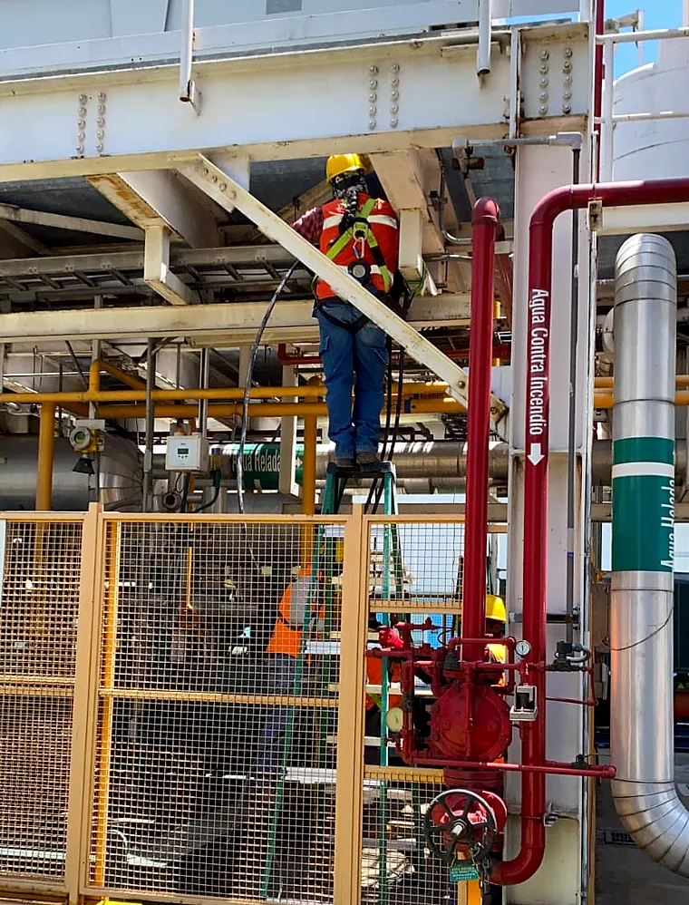 Técnico con casco, arnés y chaleco de alta visibilidad trabajando sobre una escalera bajo un rack de tubería de planta, junto a la línea roja de agua contra incendio
