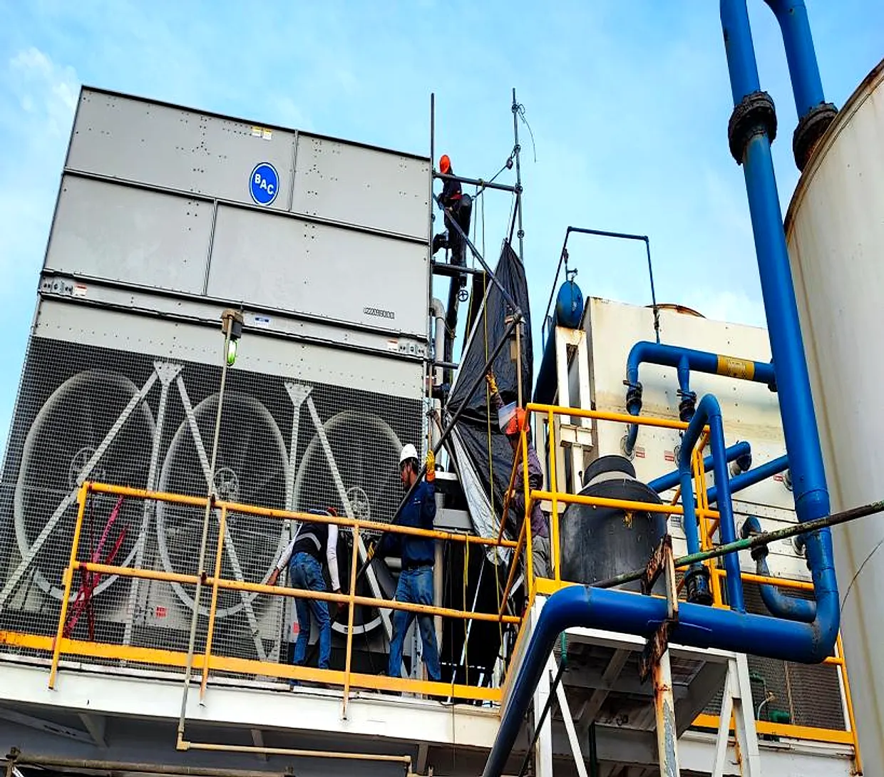 Cuadrilla con casco y arnés trabajando sobre una torre de enfriamiento, con tubería de proceso alrededor