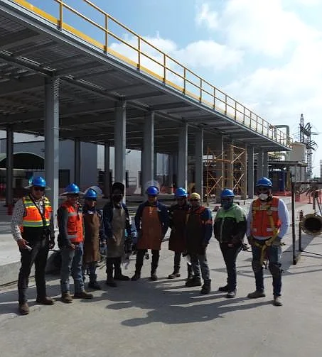 Cuadrilla de ingeniería con casco, chaleco y arnés frente a un rack de tubería
